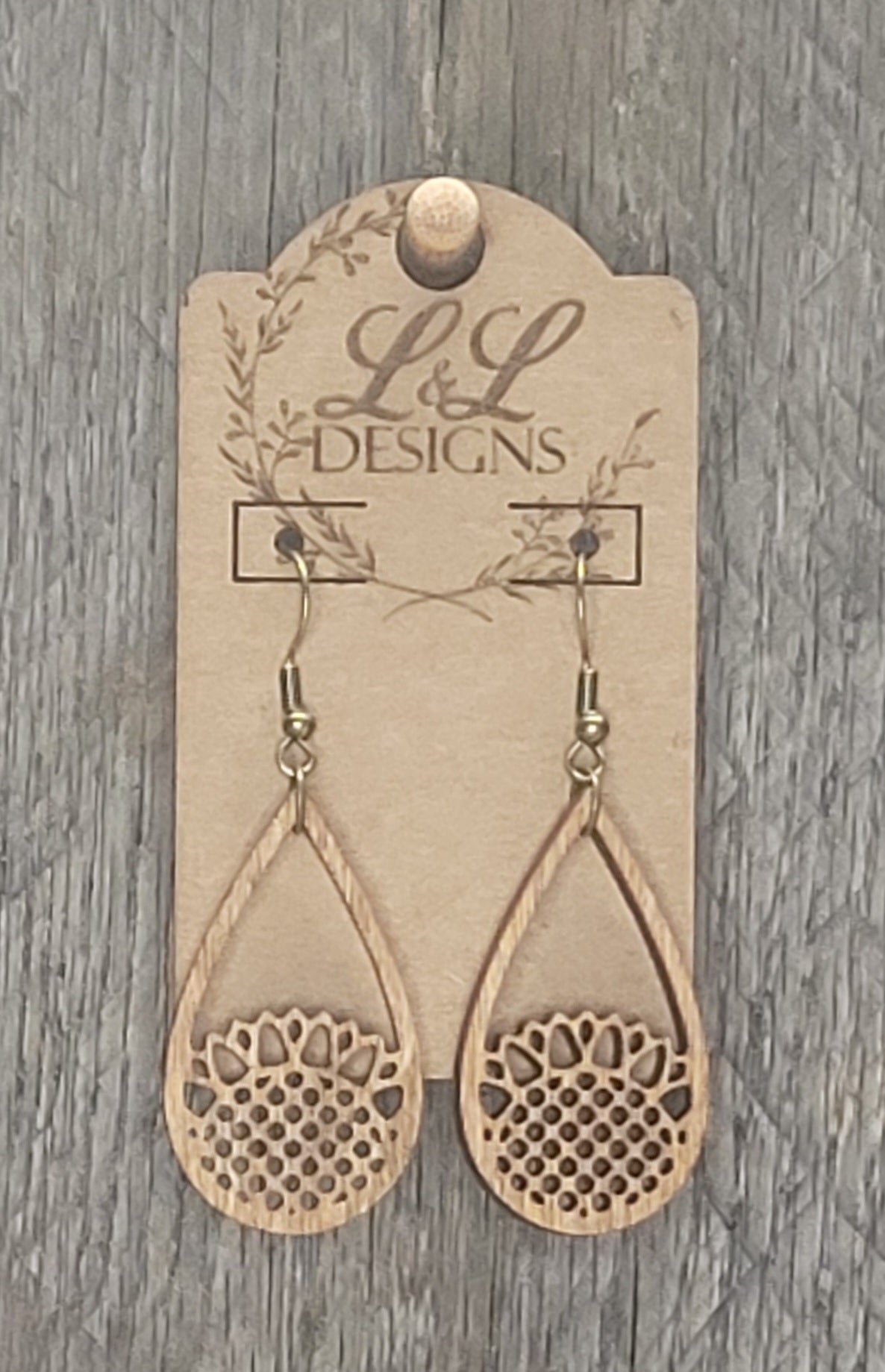 Teardrop-shaped wood earrings with intricate sunflower cutout design and dotted center displayed on L&L Designs branded card against weathered wood background.