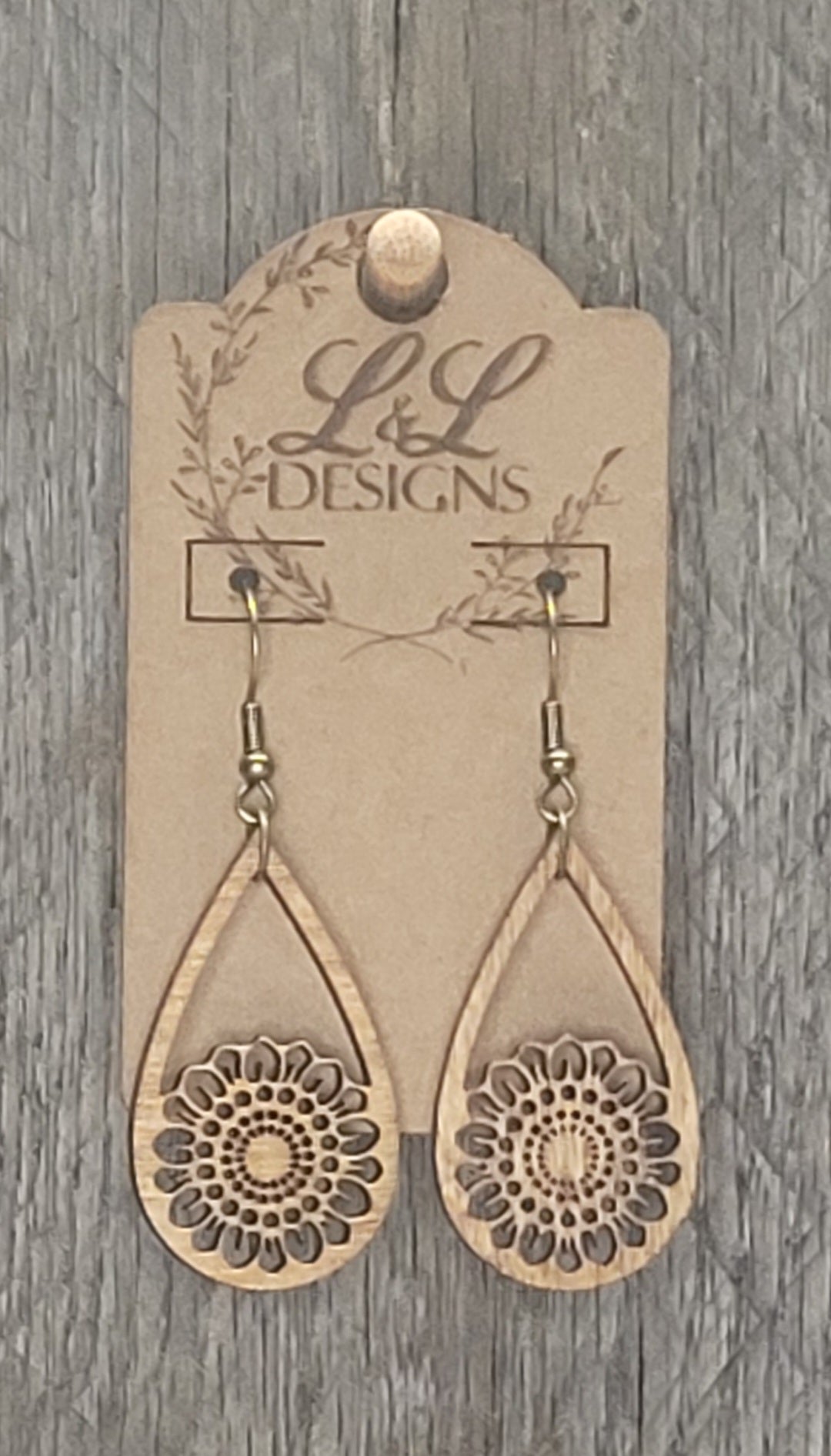 Teardrop-shaped wood earrings with circular mandala-style sunflower cutout at base displayed on L&L Designs branded card against weathered wood background