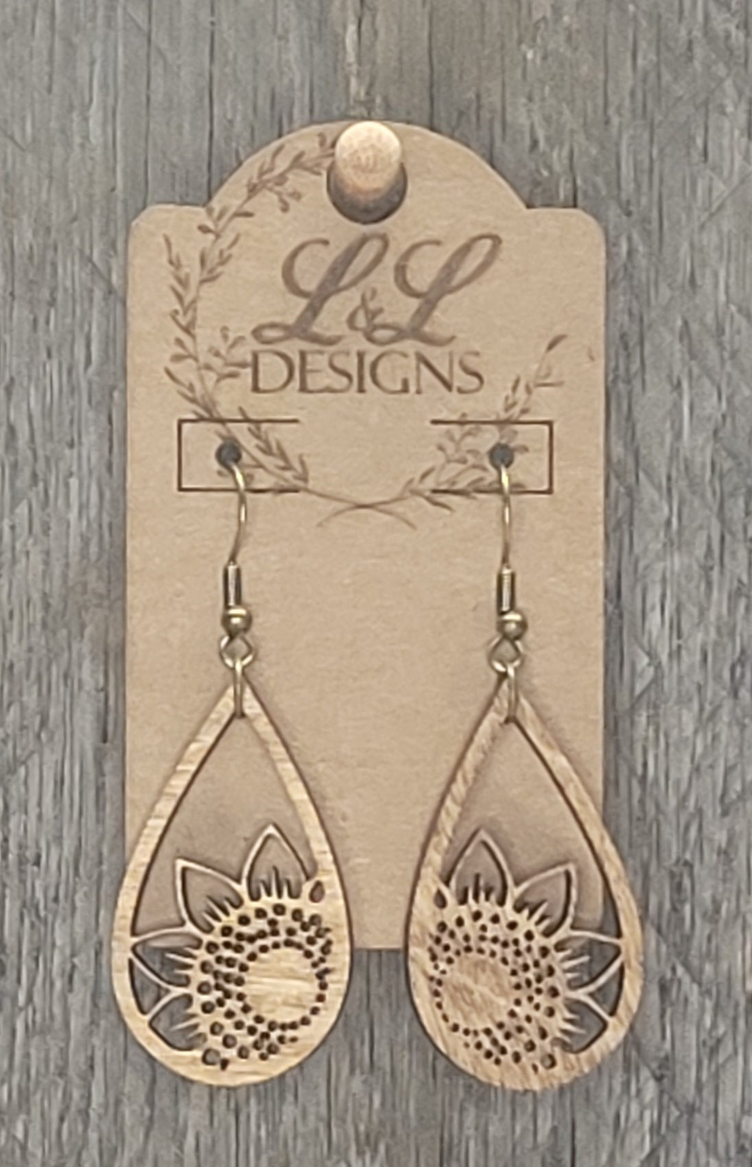 Teardrop-shaped wood earrings with sunflower and leaf cutout design at base displayed on L&L Designs branded card against weathered wood background