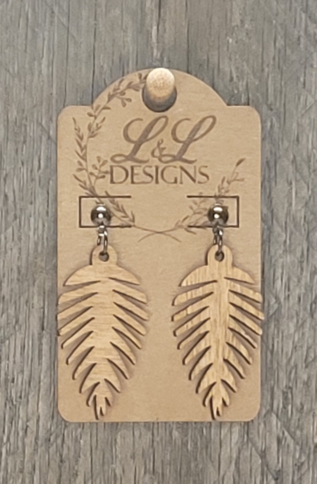 Feather-shaped wood earrings with detailed cutout lines displayed on L&L Designs branded card against weathered wood background.