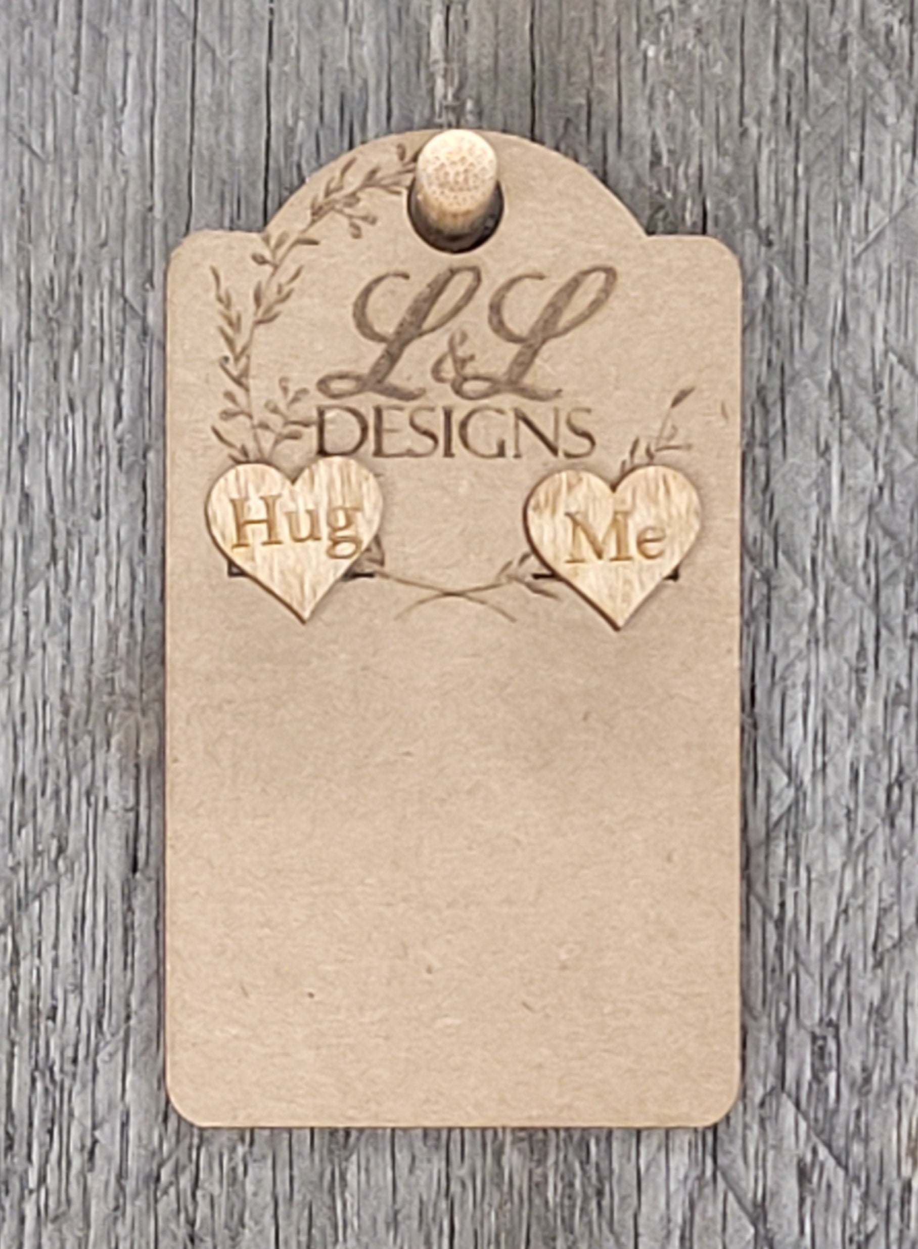 Heart-shaped wood stud earrings with 'Hug Me' engraved message displayed on L&L Designs branded card against weathered wood background.