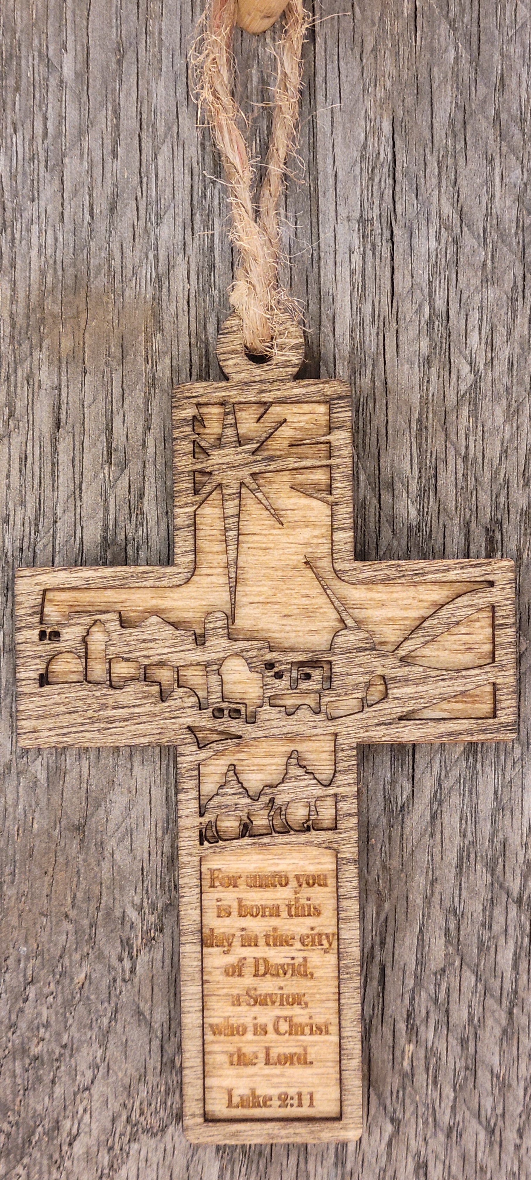 Laser-engraved wooden cross ornament featuring the Star of Bethlehem at top, Luke 2:11 scripture text reading 'For unto you is born this day in the city of David, a Savior, who is Christ the Lord,' a detailed Bethlehem cityscape across the horizontal beam, and three wise men traveling with camels below, all precision-engraved on natural wood with visible grain patterns, displayed on a wooden surface.